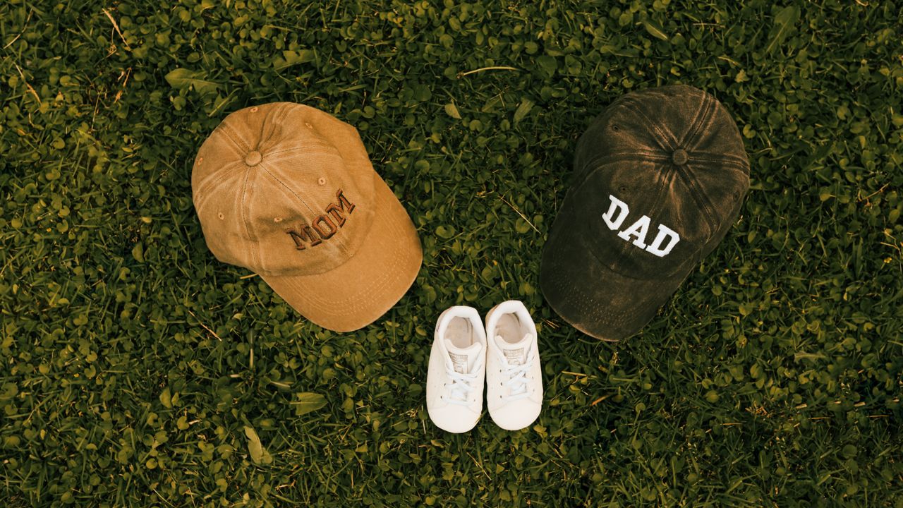 Unique family announcement concept with 'Mom', 'Dad' caps and baby shoes on grass.